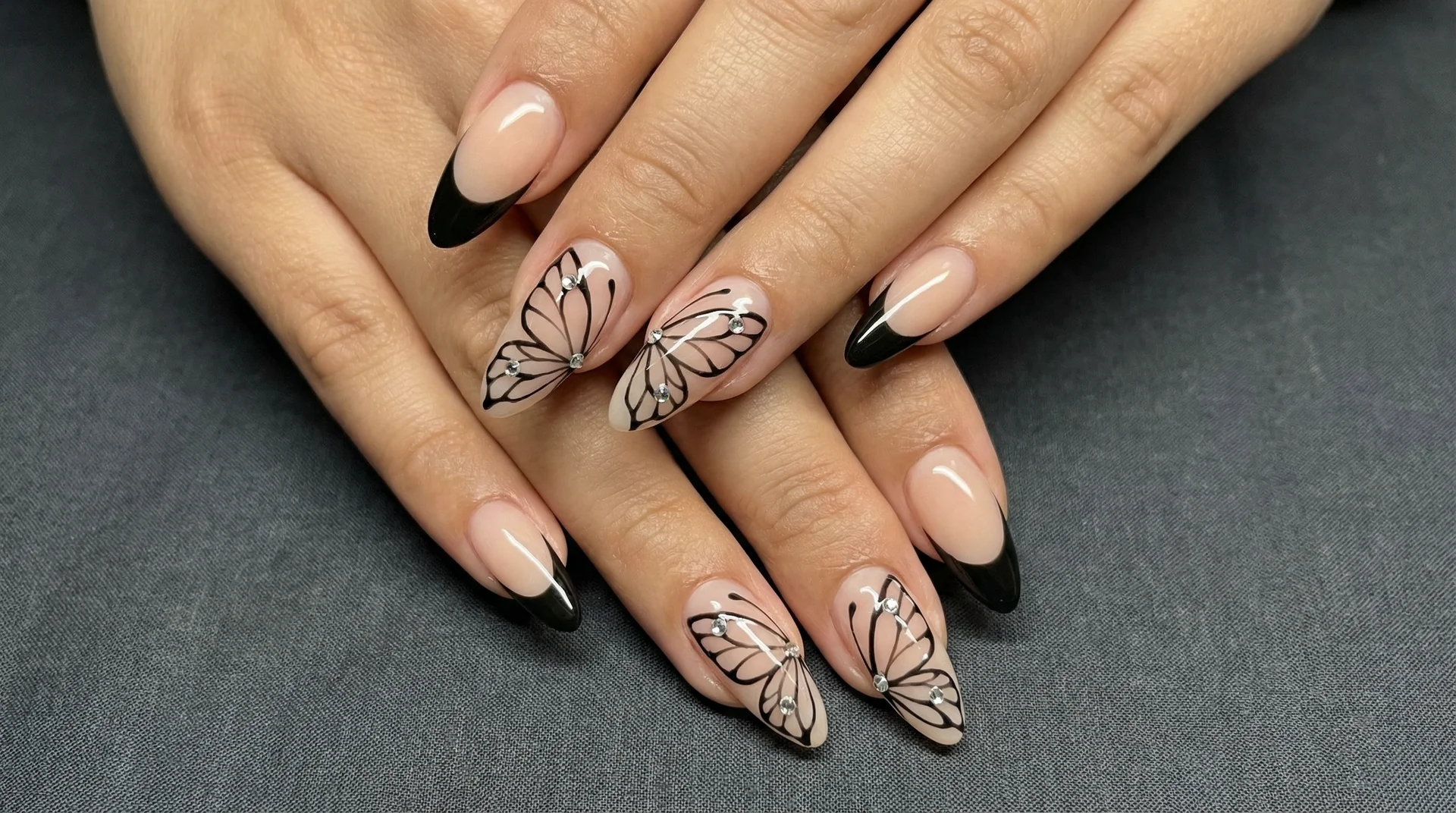 Almond nails with black butterfly wing tip line art and crystal accents on nude base