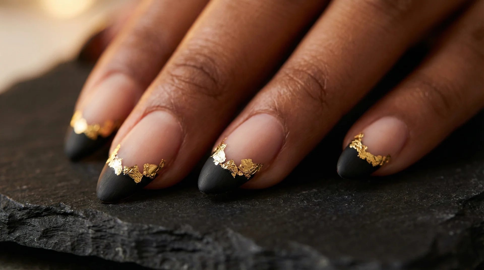 Almond nails with black french tip edged in irregular gold foil flakes on deep warm skin