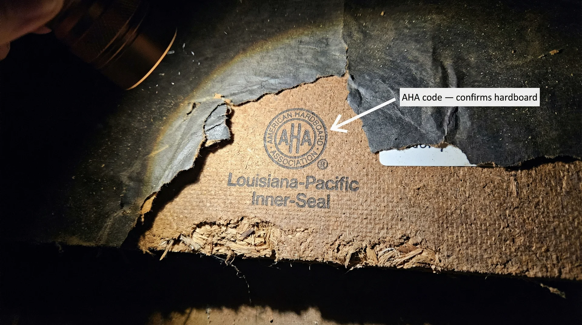 Back of hardboard siding panel in attic showing AHA stamp and wood fiber texture along cut edge
