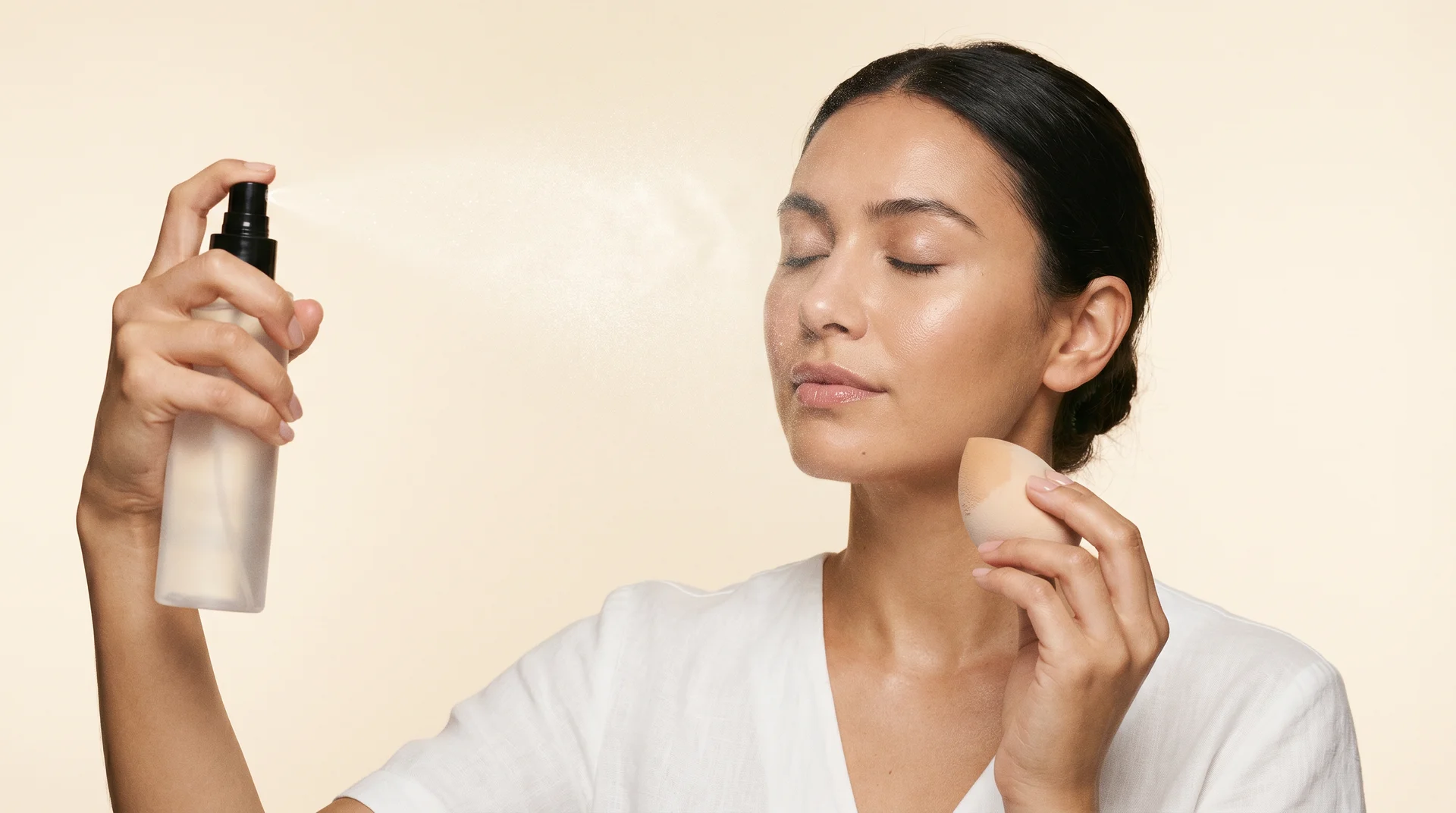 Woman misting setting spray at arm's length with a damp beauty sponge in hand ready to press and fix patchy foundation