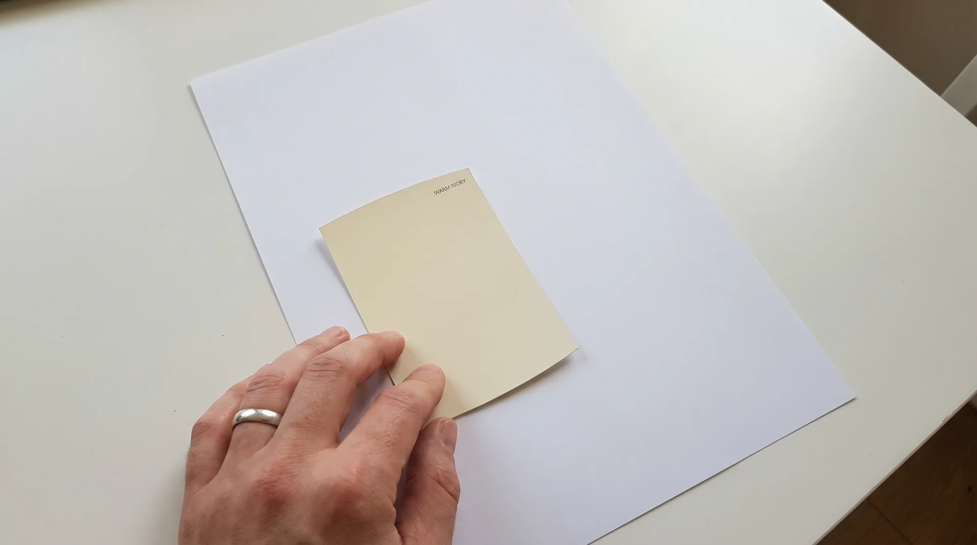 Hand holding an off white paint chip against white printer paper to reveal yellow undertone by comparison